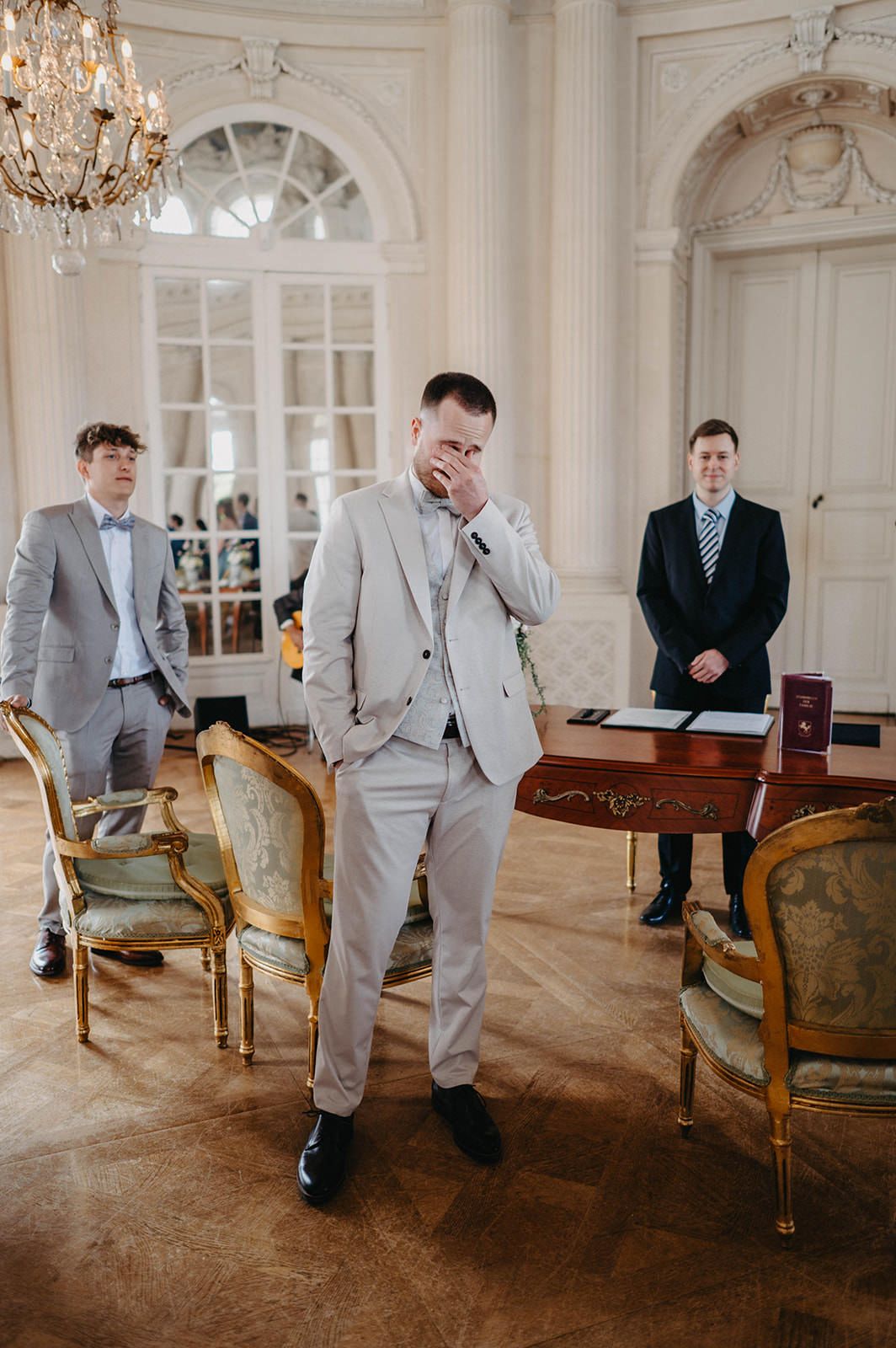 Elegant wedding scene in ornate room with chandelier, antique furniture and three men in formal attire.