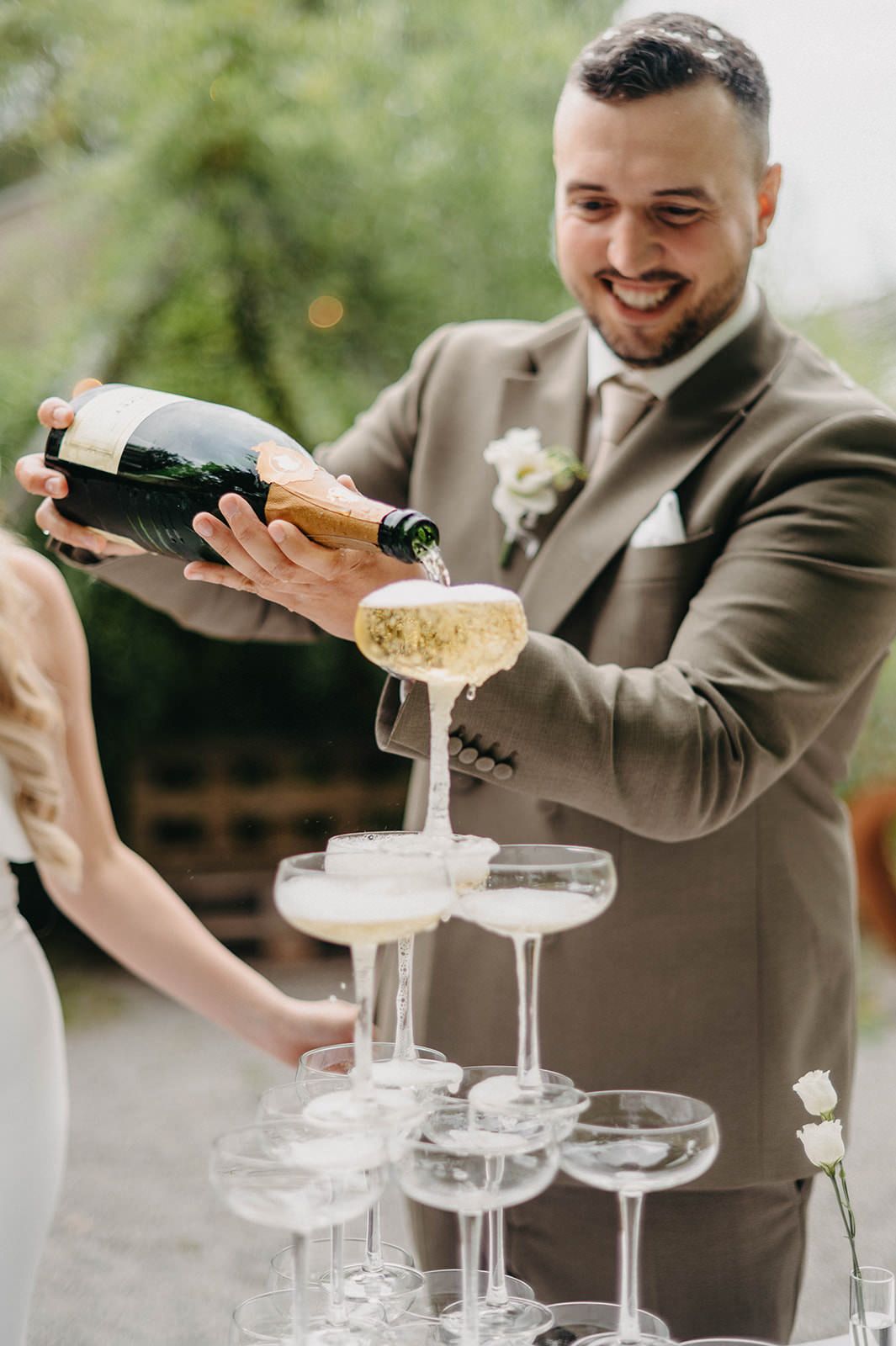 Person in brown suit pouring champagne into a tower of coupe glasses at an elegant outdoor celebration.