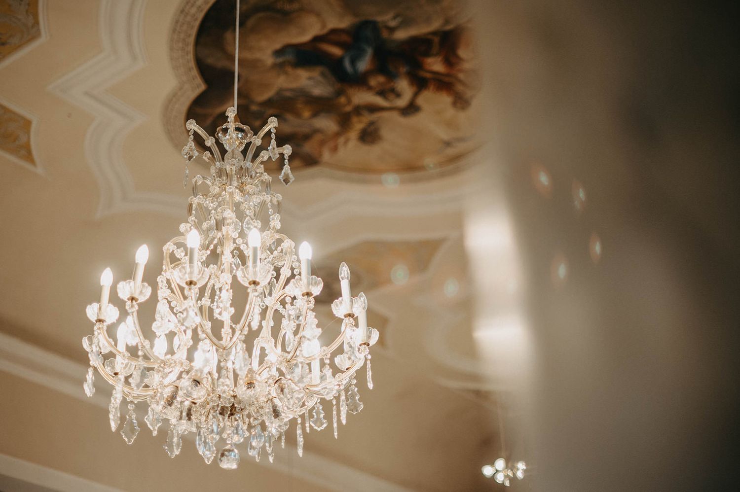 Elegant crystal chandelier hanging from ornate ceiling with decorative painted details creates luxurious ambiance.