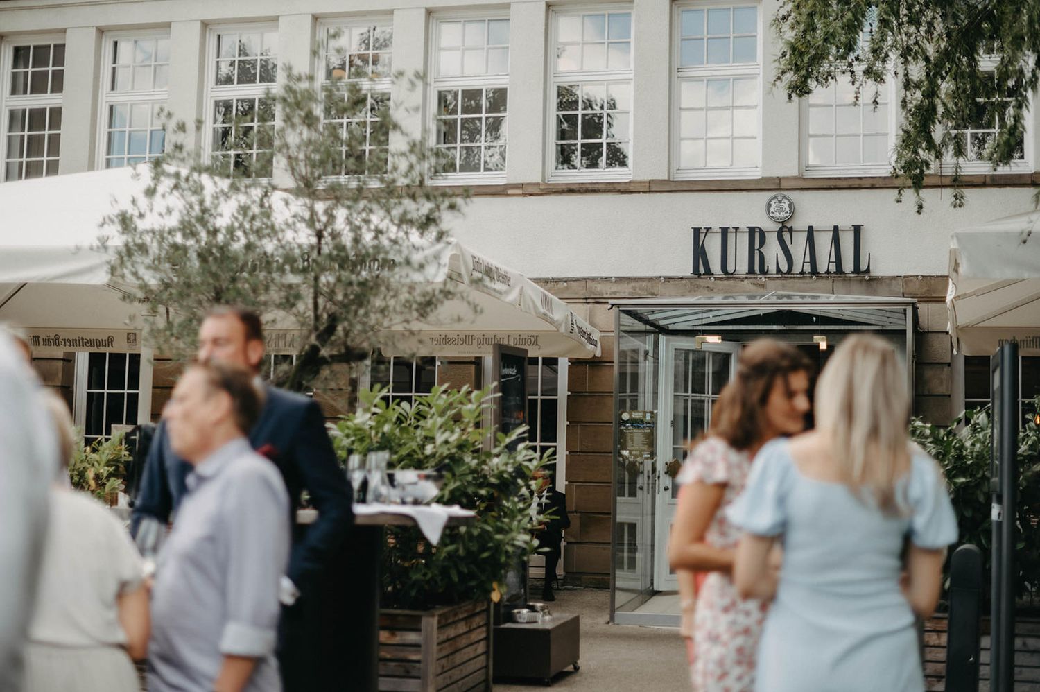 Wedding guests mingle and socialize on an outdoor patio near a building with Kursaal signage.