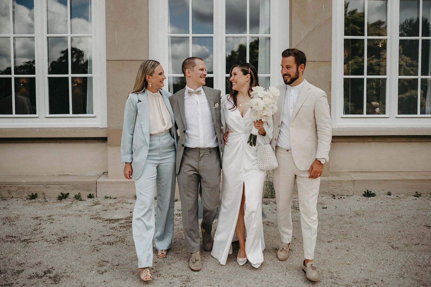 Wedding party in light blue and white attire walks together outside elegant venue with white columns and windows.