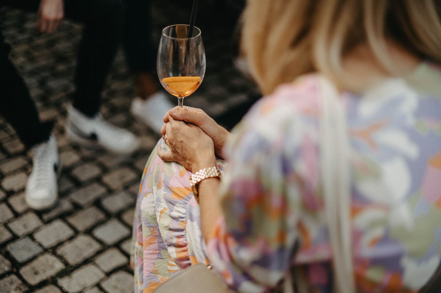 People holding wine glasses with orange-tinted beverages during an outdoor gathering on cobblestone pavement.