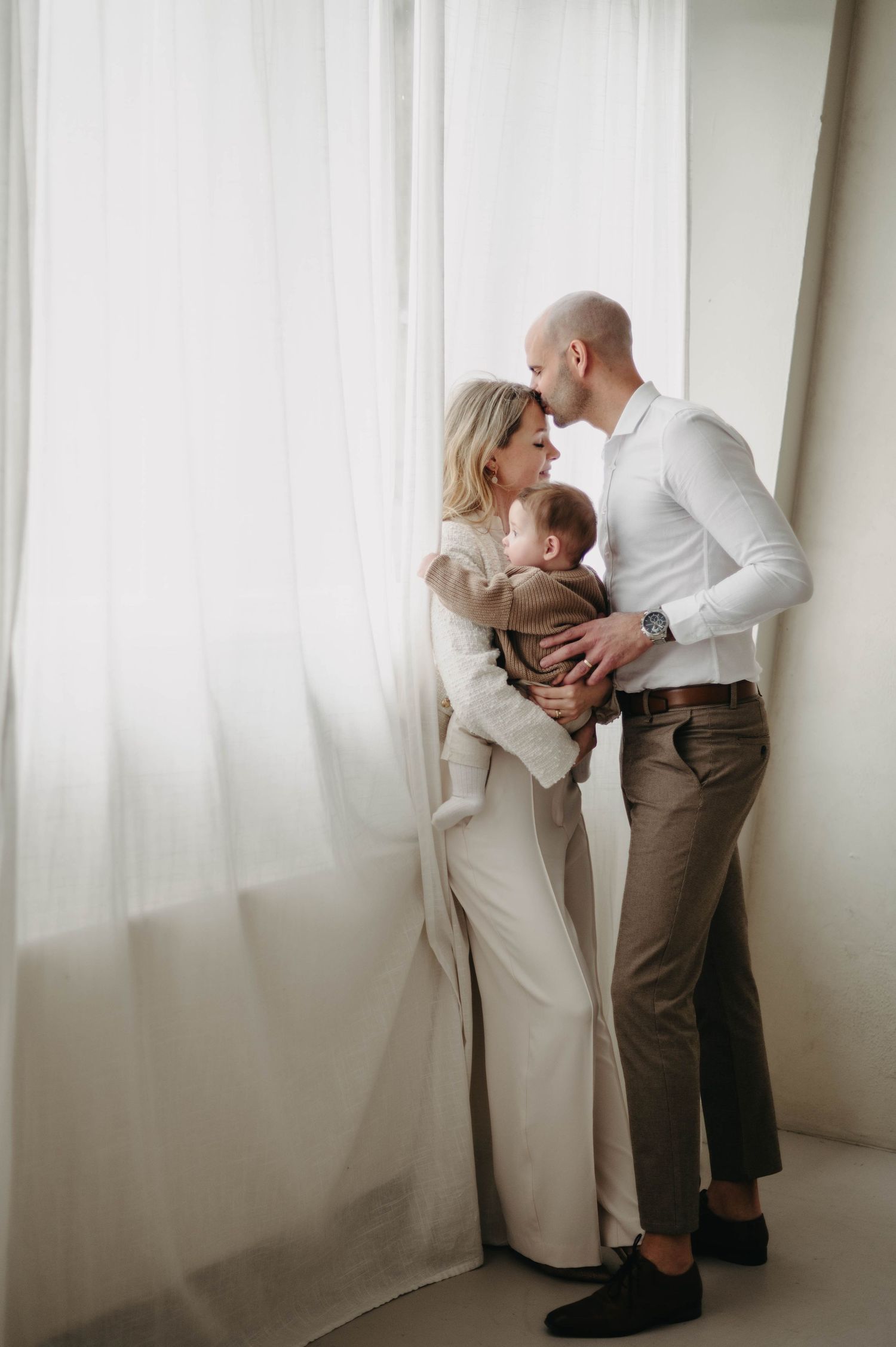 A tender family moment captured in soft light as parents embrace their baby near a white curtained window.