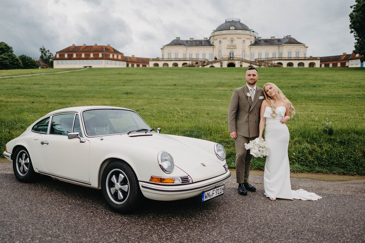 Classic white Porsche 911 parked in front of Solitude Palace in Stuttgart with couple standing beside the vintage car.