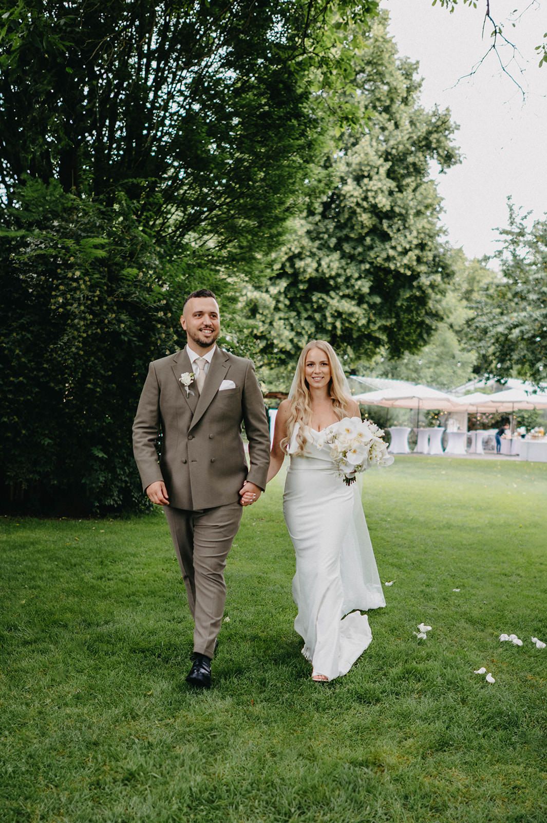 A couple in wedding attire walks together on a grassy lawn with trees in the background during an outdoor celebration.
