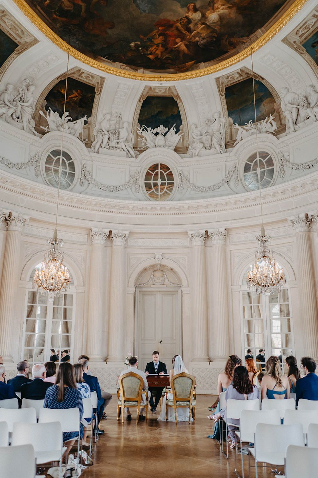 Elegant white baroque ballroom with ornate ceiling frescoes, crystal chandeliers and white chairs set up for an event.