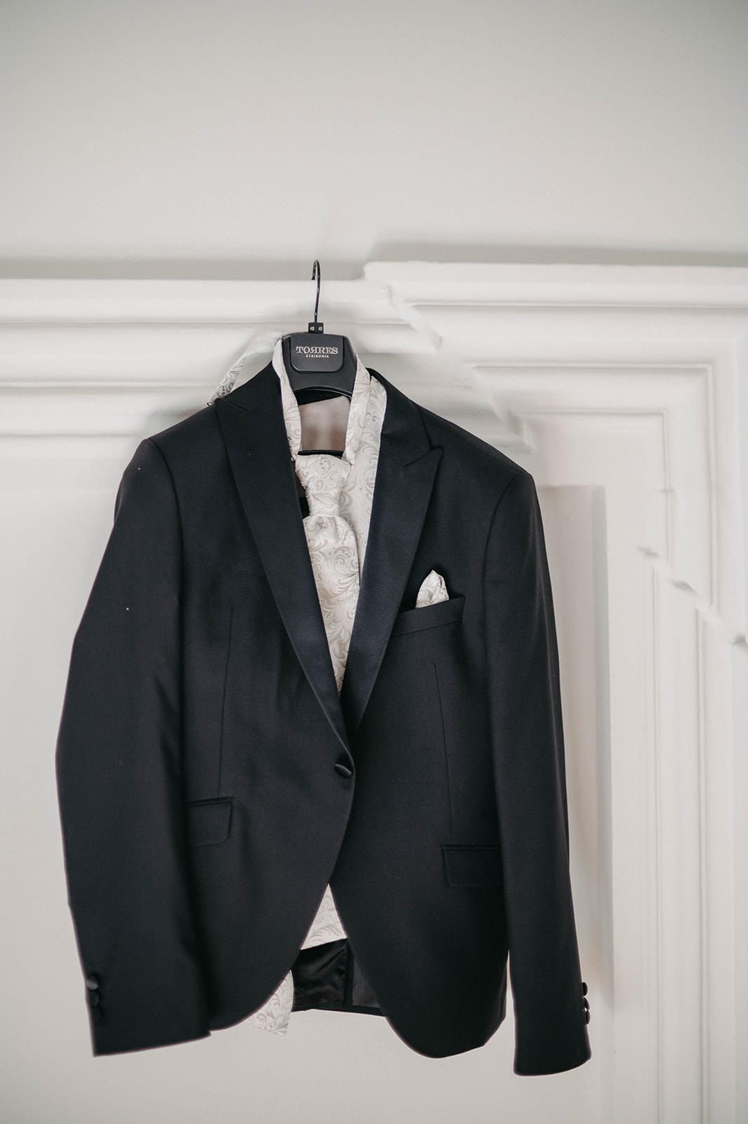 Black formal suit jackets hanging on white wall hooks display elegant wedding attire with white pocket squares.