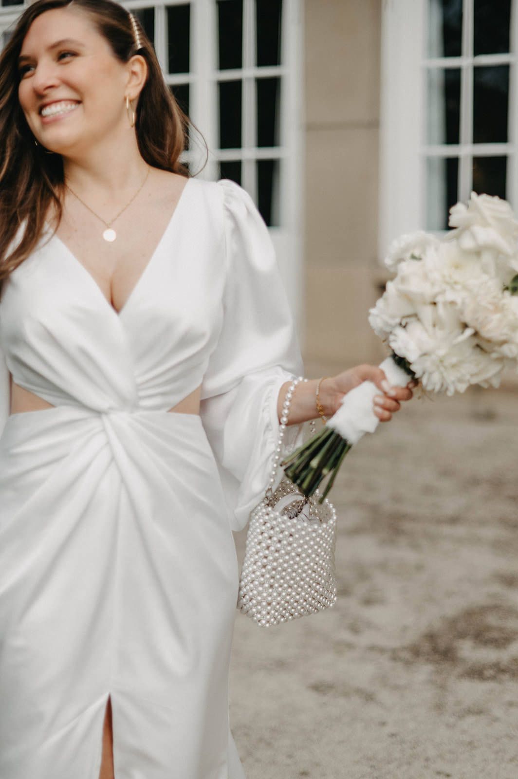 Elegant bride in white long-sleeved dress holds a sparkling silver handbag and white floral bouquet.