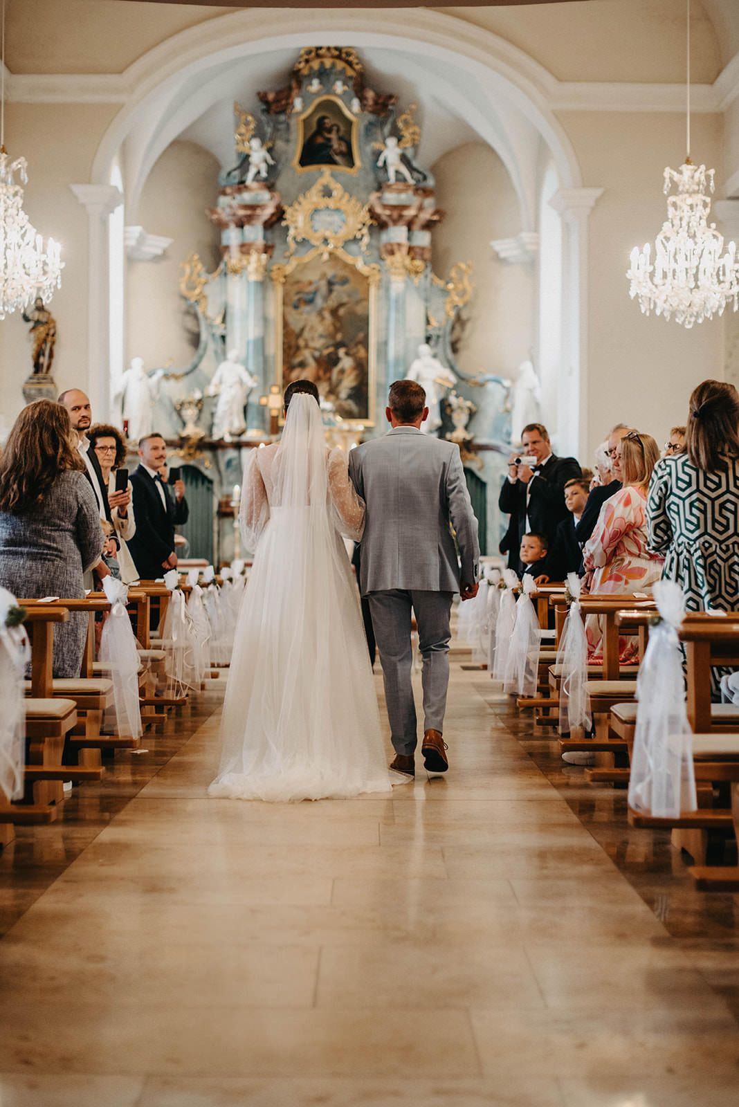A panoramic view of an elegant church wedding ceremony with guests seated in wooden pews and ornate baroque decor.
