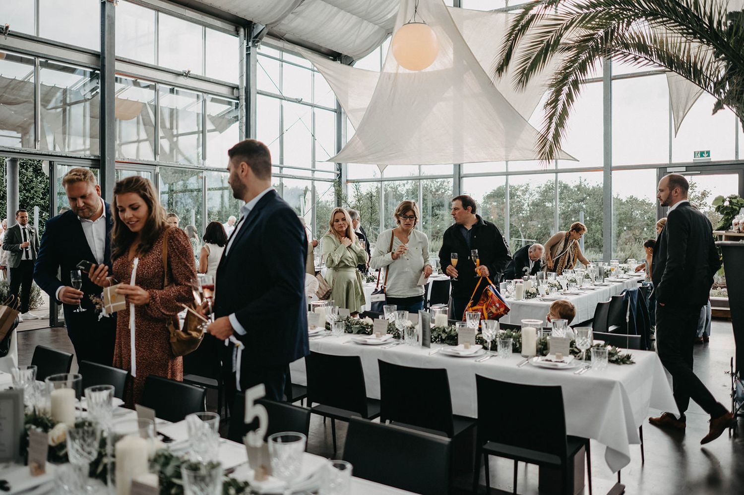 A panoramic view of guests mingling at a modern event space with floor-to-ceiling windows and palm trees outside.
