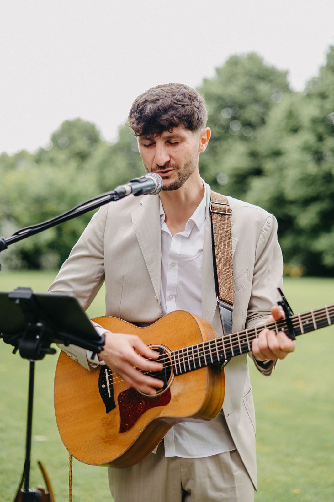 Musician in beige suit plays acoustic guitar outdoors in grassy field during summer event.