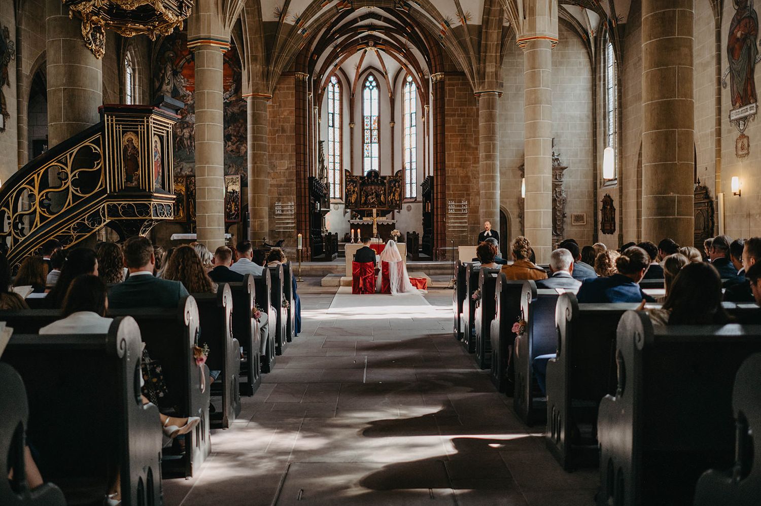 Religious ceremony taking place inside a historic Gothic church with stone arches and wooden pews filled with attendees.