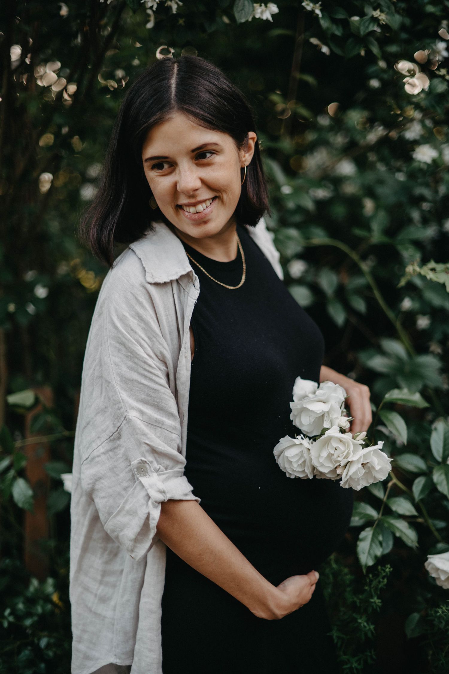 A maternity photo showing a woman in black dress holding white roses in a garden setting.