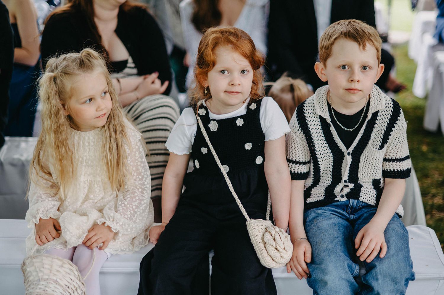 Children sit together at an outdoor event wearing casual clothing and formal attire.