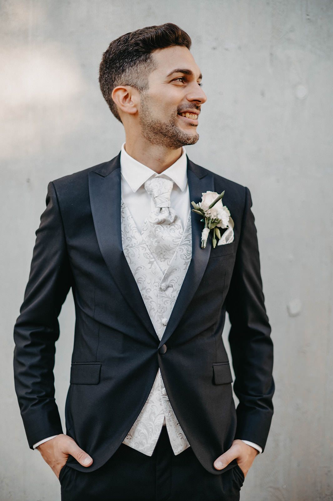 A groom in a classic black tuxedo with white vest and tie poses against a gray wall with a boutonniere.