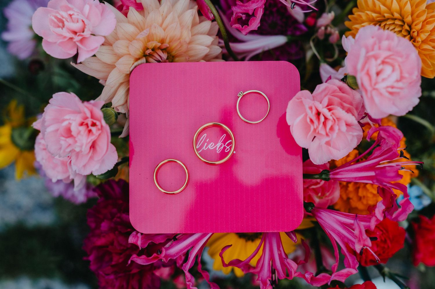Wedding rings displayed on vibrant pink cards surrounded by pink and orange flowers and floral arrangements.