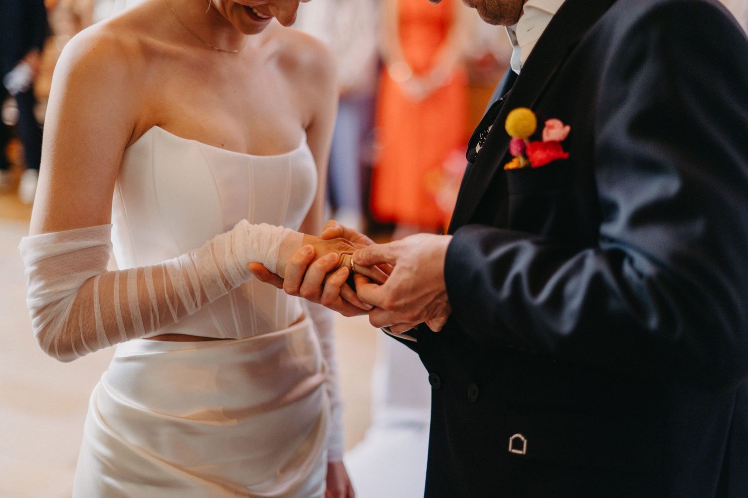A couple holds hands during an intimate wedding ceremony with orange floral decor in the background.