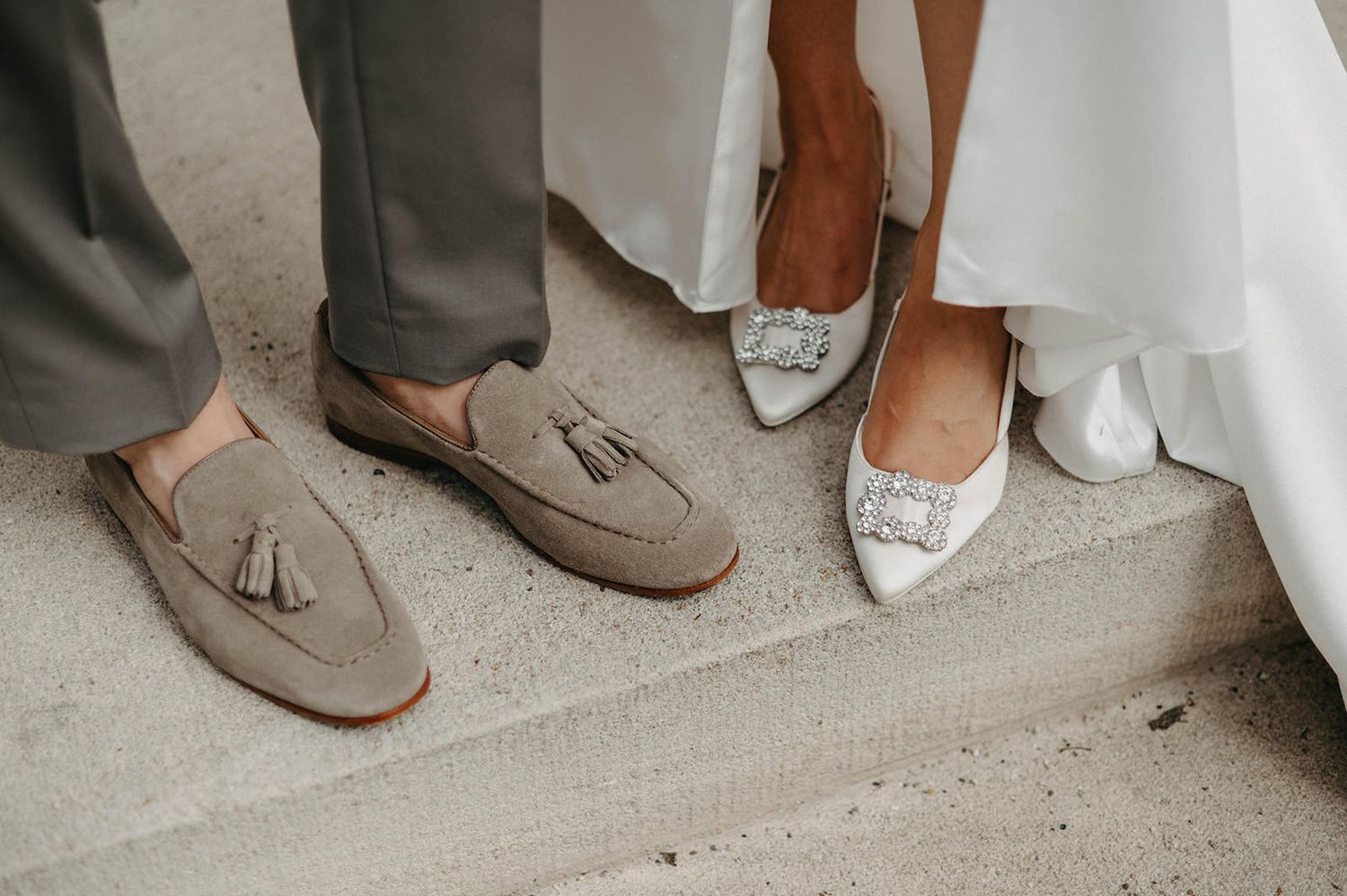 Close-up of bride and groom's wedding shoes, with grey suede loafers and white embellished bridal flats.