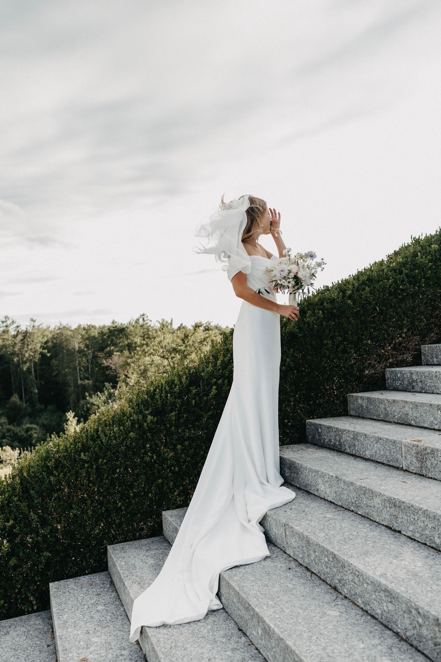 Bride in flowing white dress posing on stone steps with flowing veil in natural light.