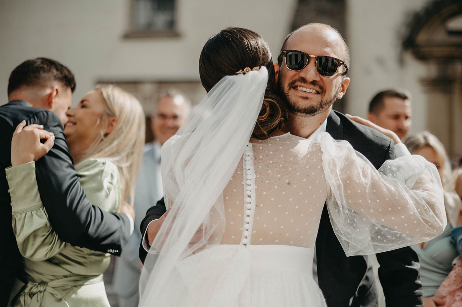 Wedding guests celebrate and embrace during an outdoor reception on a sunny day.
