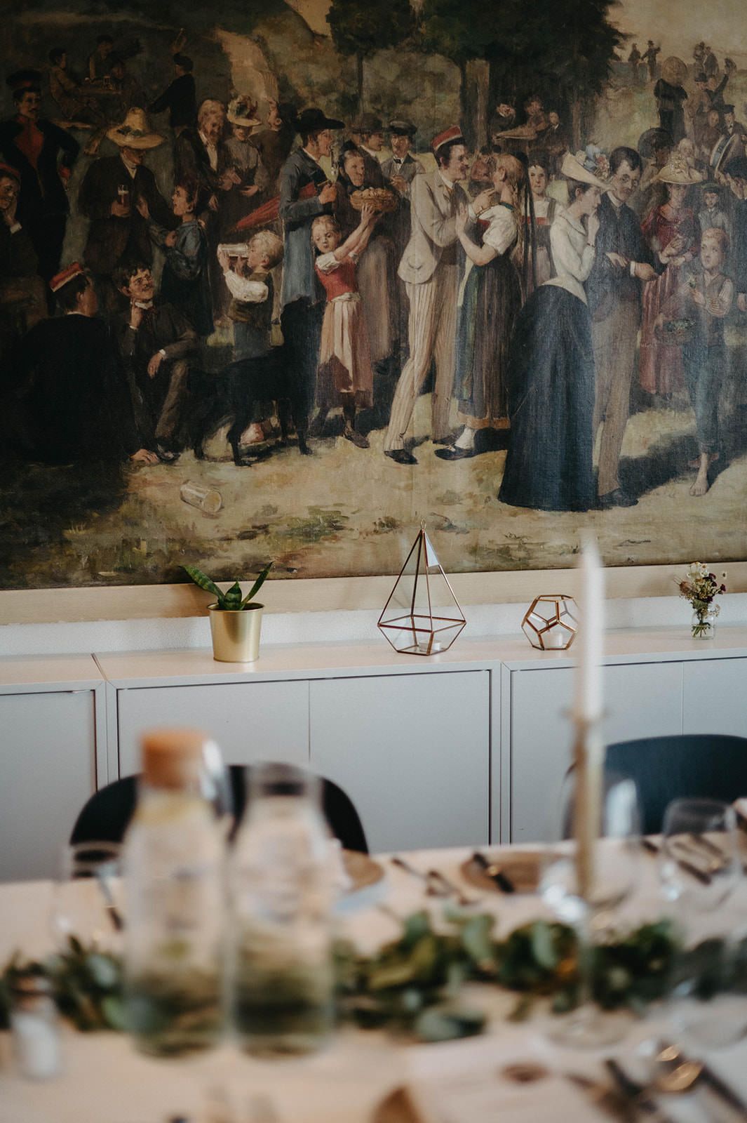 Rustic dining room interior with vintage mural artwork on wall above white cabinets and place settings on table.