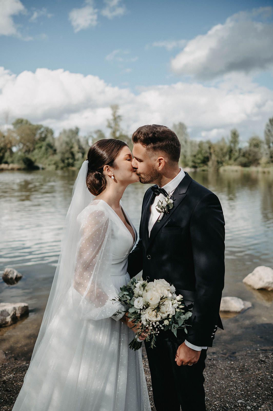 Romantic wedding couple embraces by a serene lake with white florals and a flowing veil.