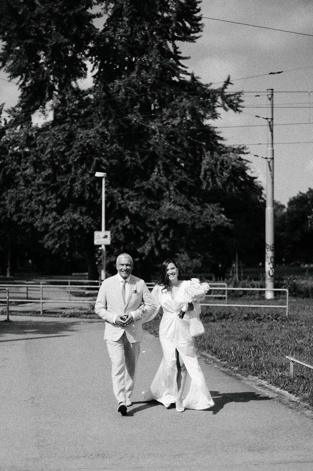 A couple in white outfits walks along a path near trees and utility poles in a vintage black and white photograph.