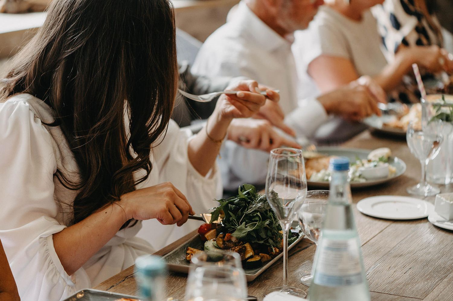 Diners enjoy a meal at a table with wine glasses and fresh greens on white linens.