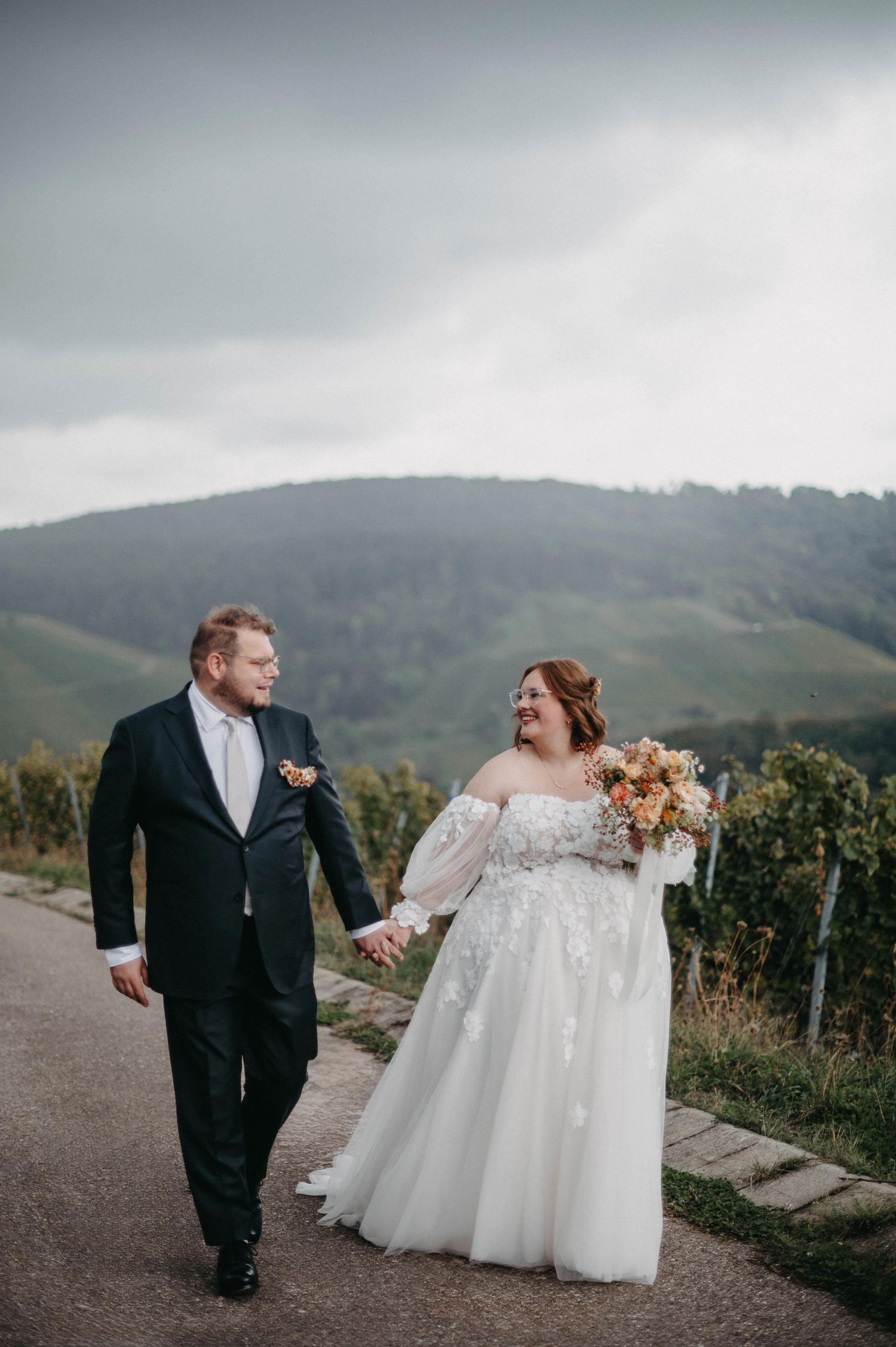A couple shares candid moments on a hilltop path surrounded by misty mountains and vineyards.