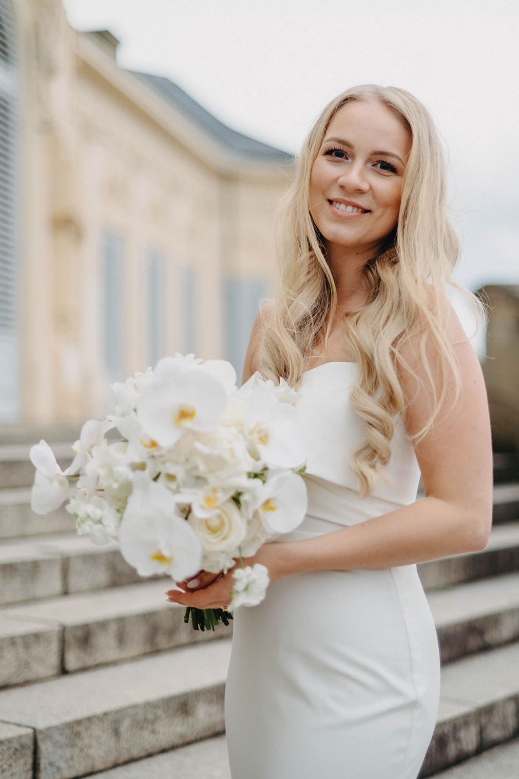 Elegant bride in white dress holding beautiful white orchids on a grand balcony.