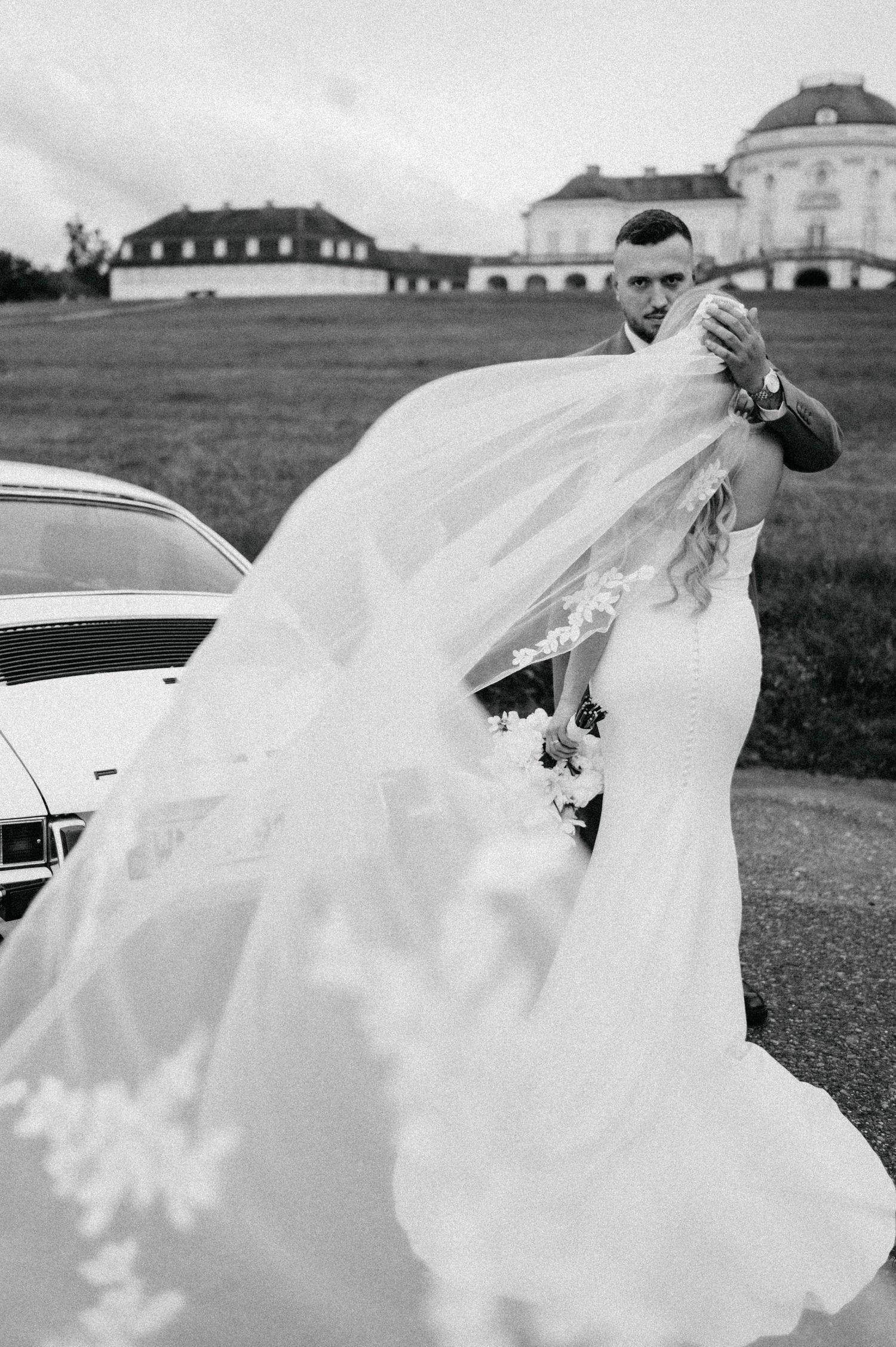 Wedding couple with flowing veil caught in wind outside historic building.