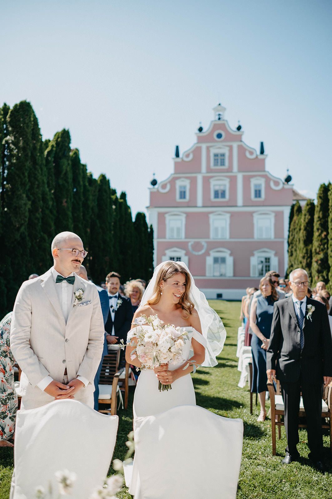 Outdoor wedding ceremony in front of a pink baroque-style building with guests gathered on a sunny lawn.