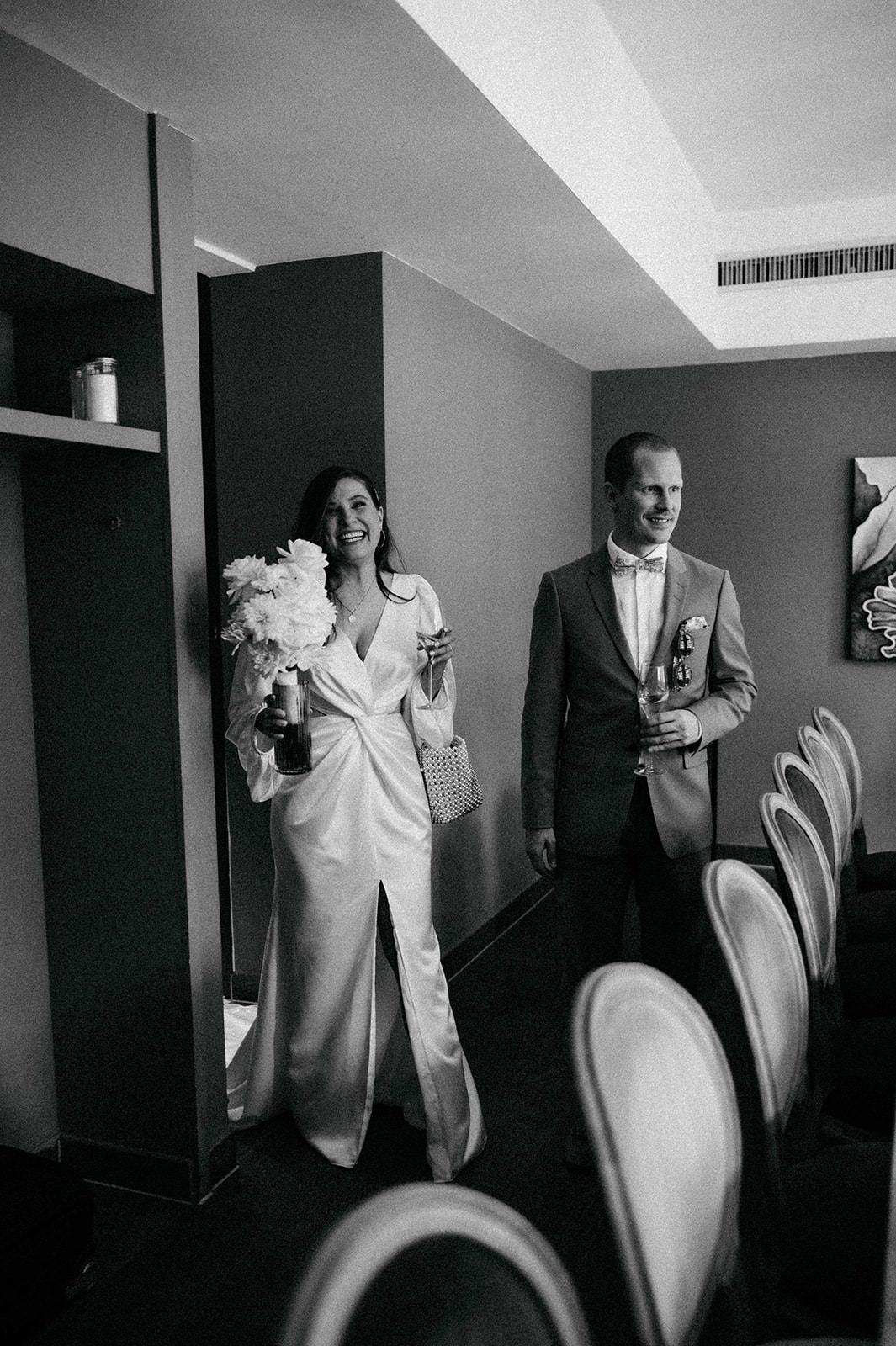 Black and white photo of wedding party members in formal attire holding coffee cups in a hotel hallway.