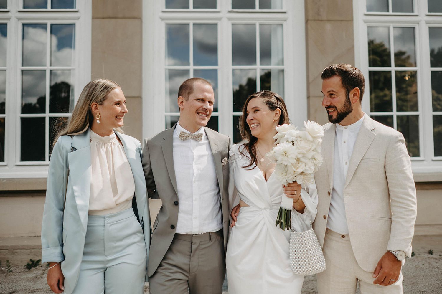 Group of wedding attendees sharing joyful moment outside elegant venue with white columns.