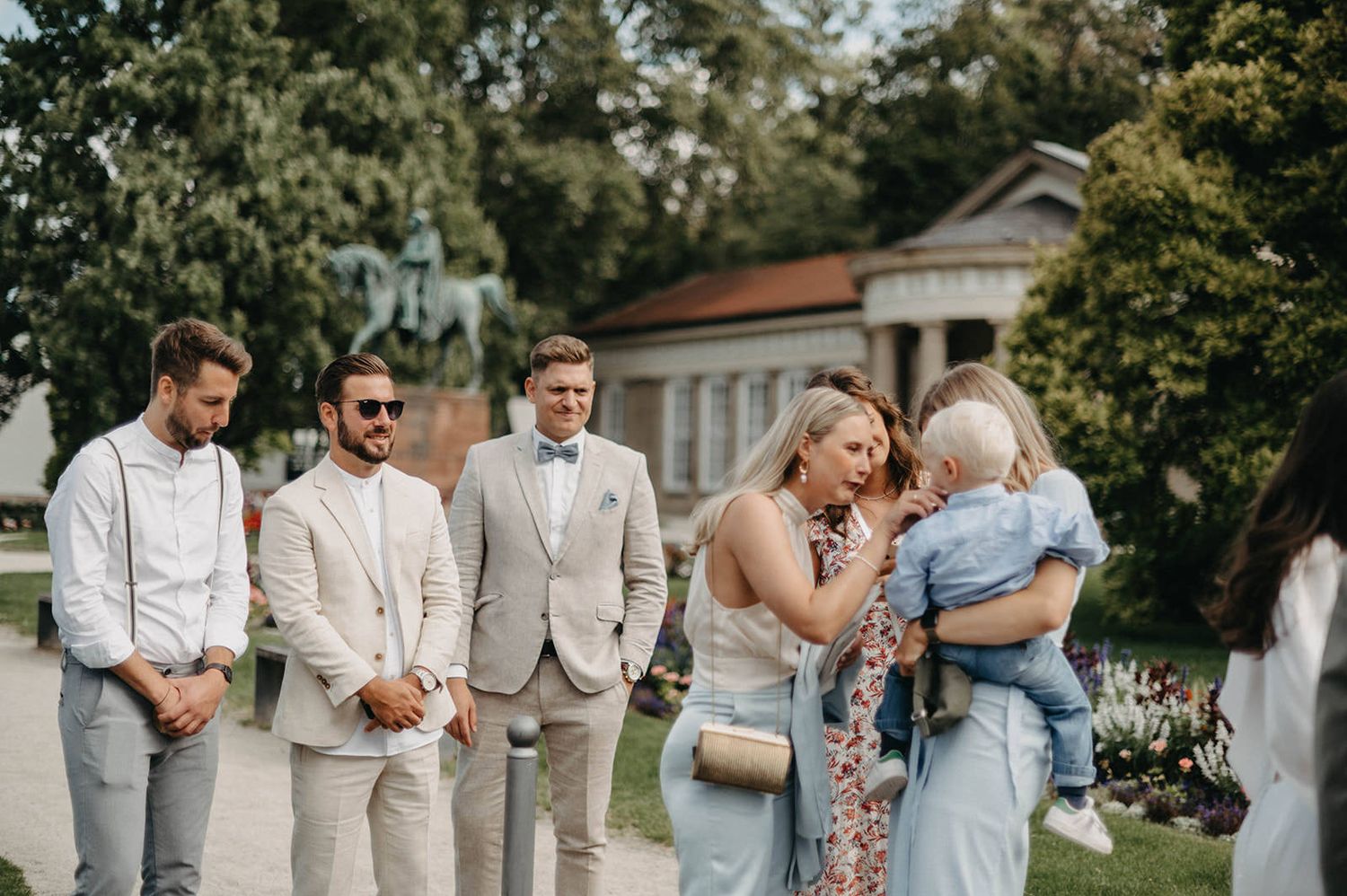 Wedding party members gather outdoors in light summer attire on a sunny day with a white gazebo in the background.