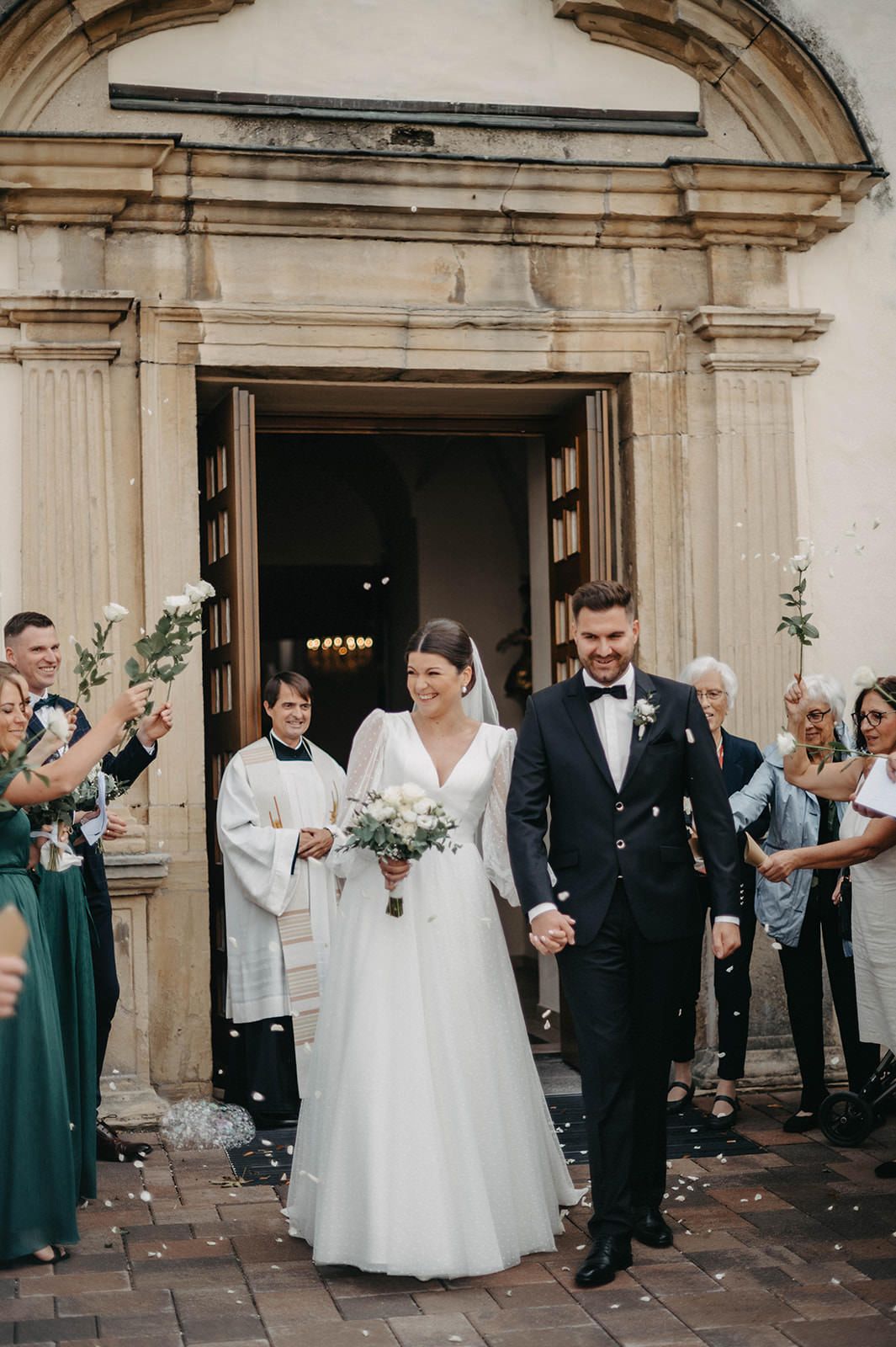 Wedding party celebrates with confetti toss outside historic stone church entrance during joyful recessional.