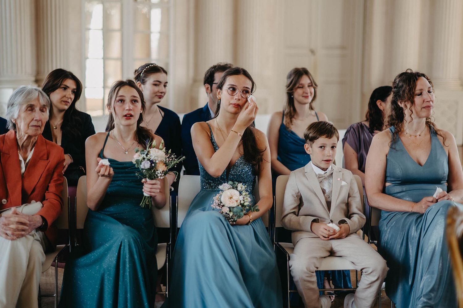 Wedding guests in blue dresses and formal attire watch emotionally during ceremony.