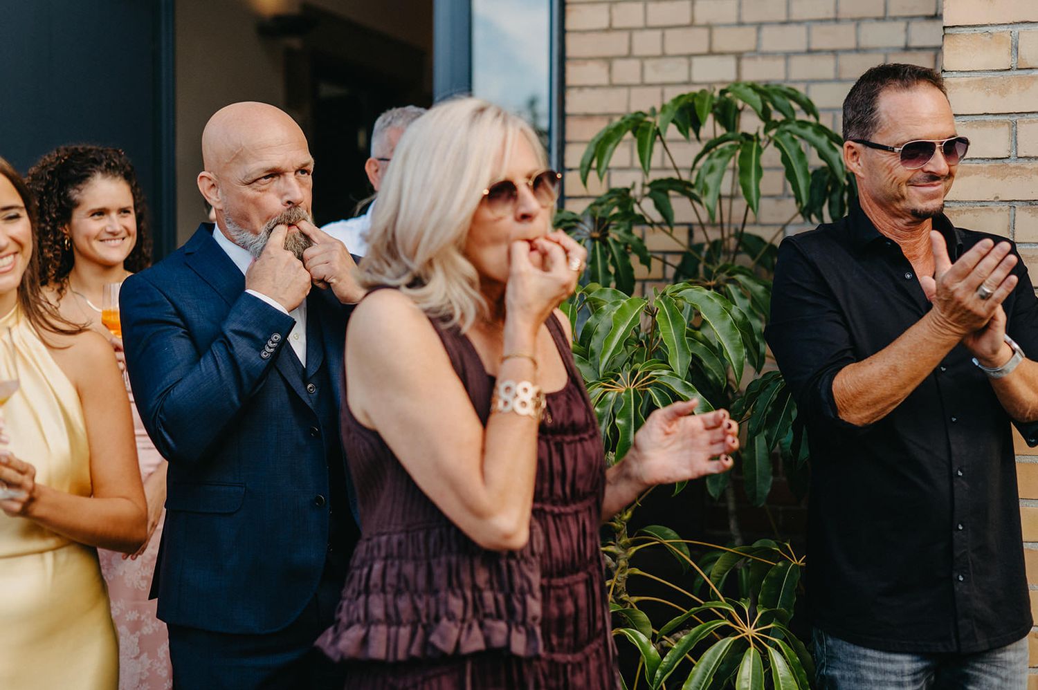 Wedding guests stand in a line outdoors at a celebration, wearing formal attire and enjoying the festivities.