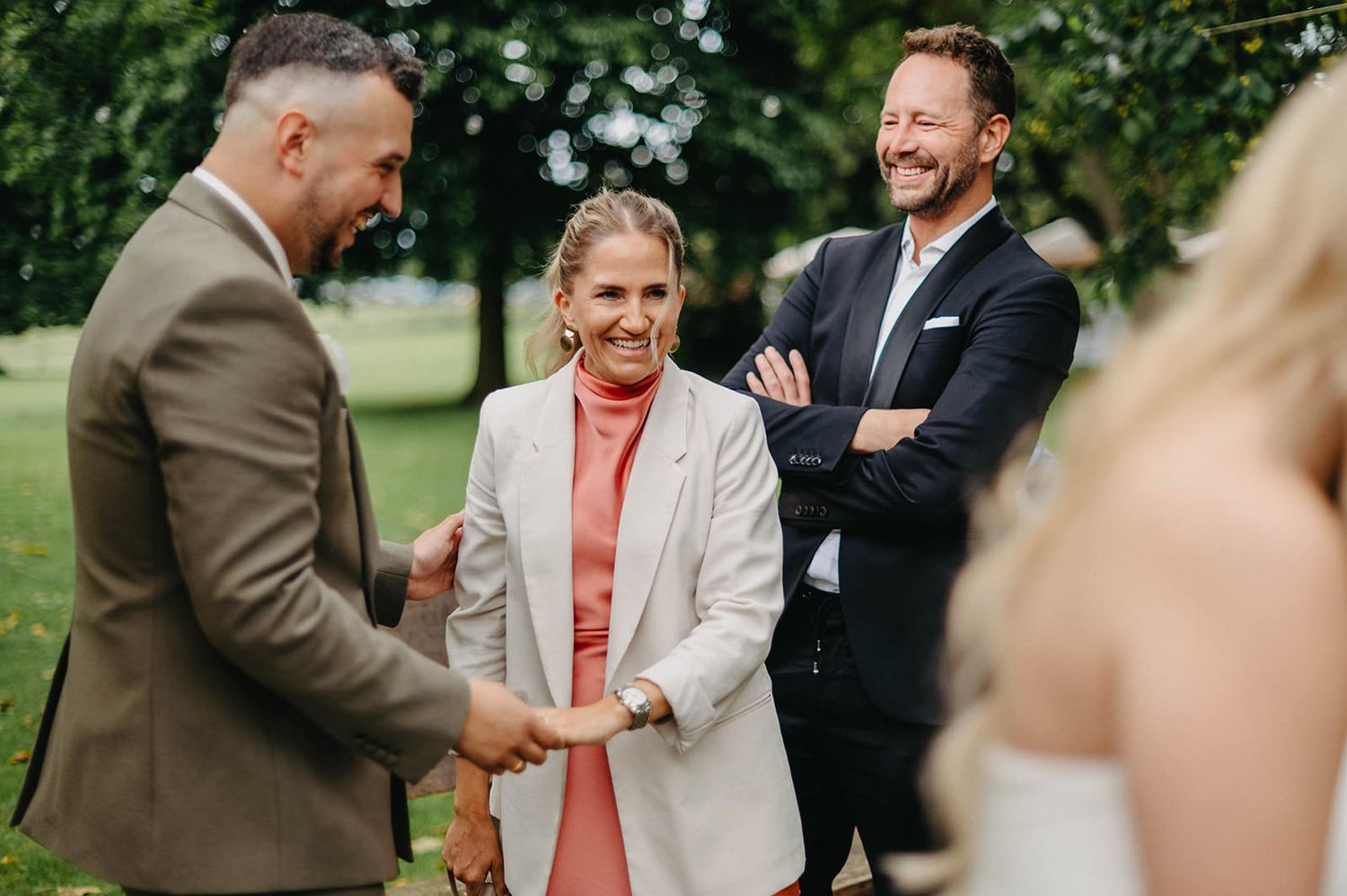 A group of well-dressed people share a cheerful moment outdoors on a sunny day in a garden setting.