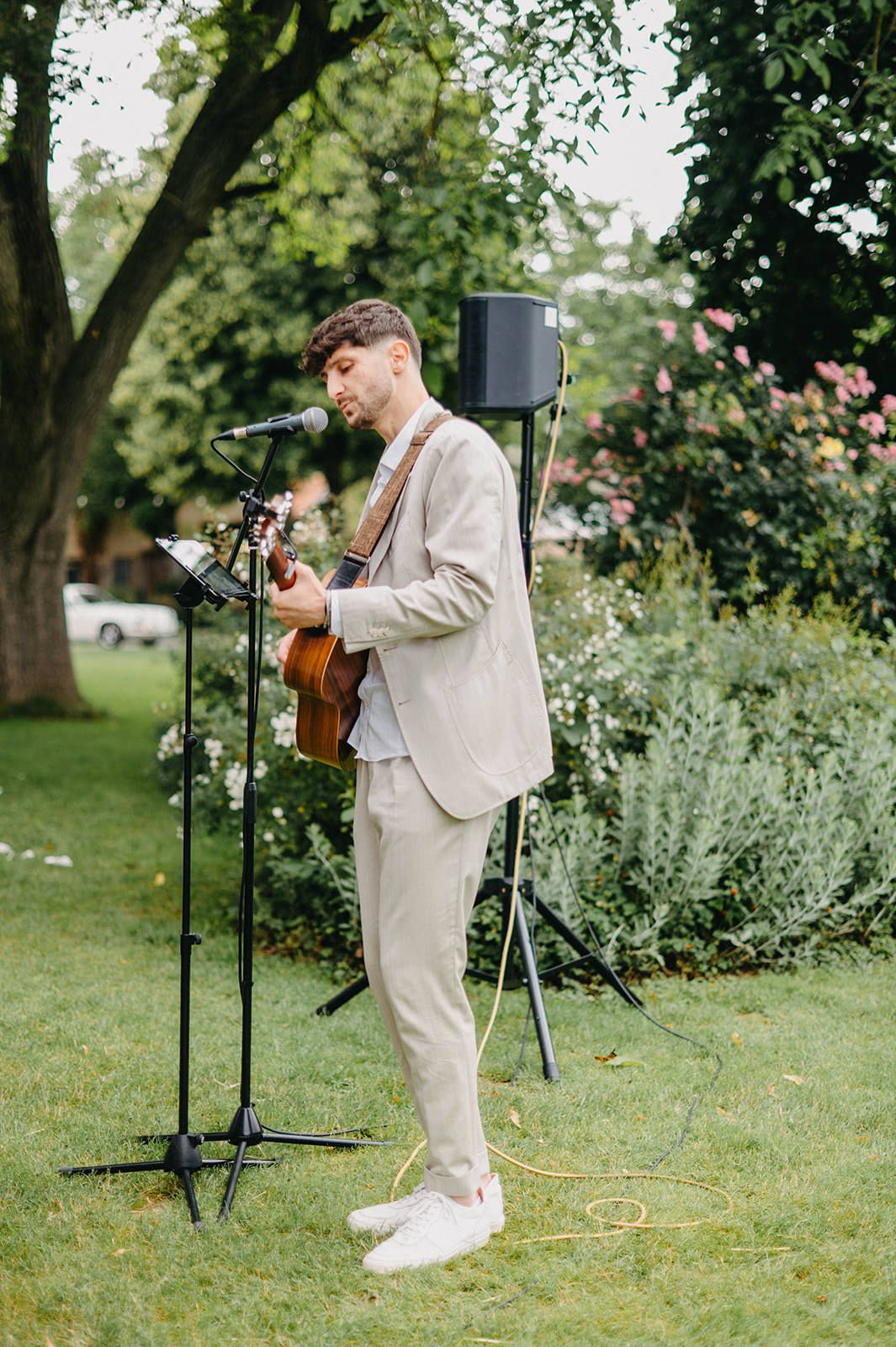 A musician in a light colored suit performs outdoors in a garden setting with microphone stands and equipment.