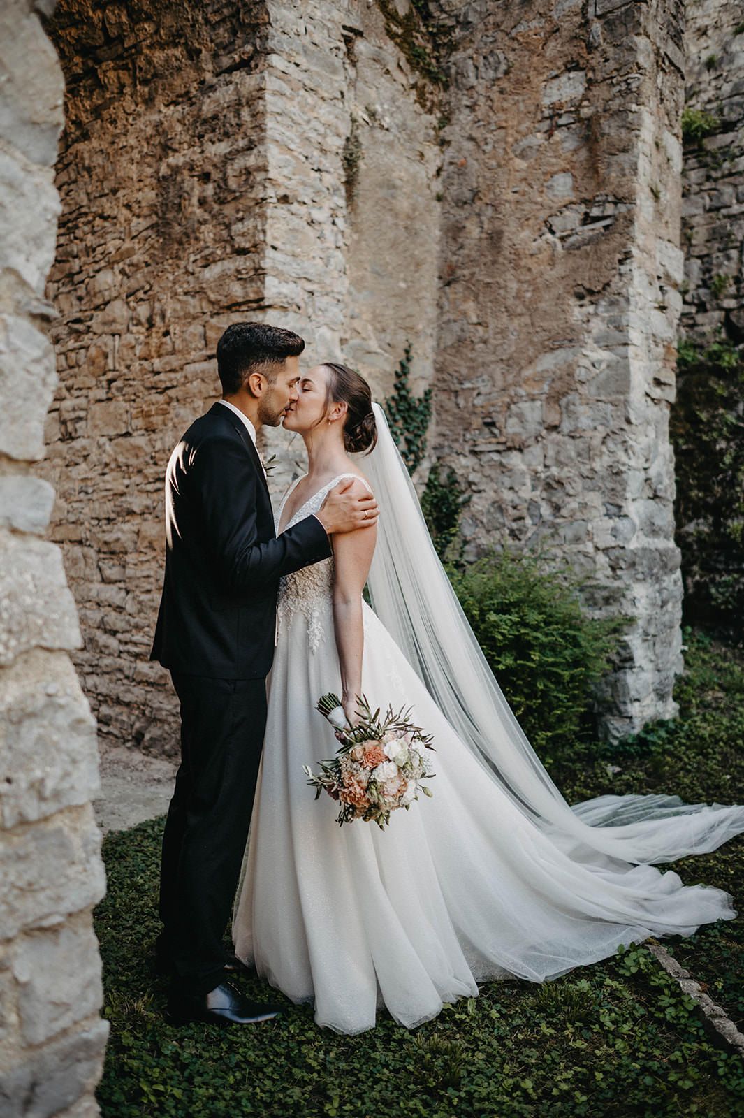 A romantic wedding couple shares a tender moment among ancient stone columns and greenery.