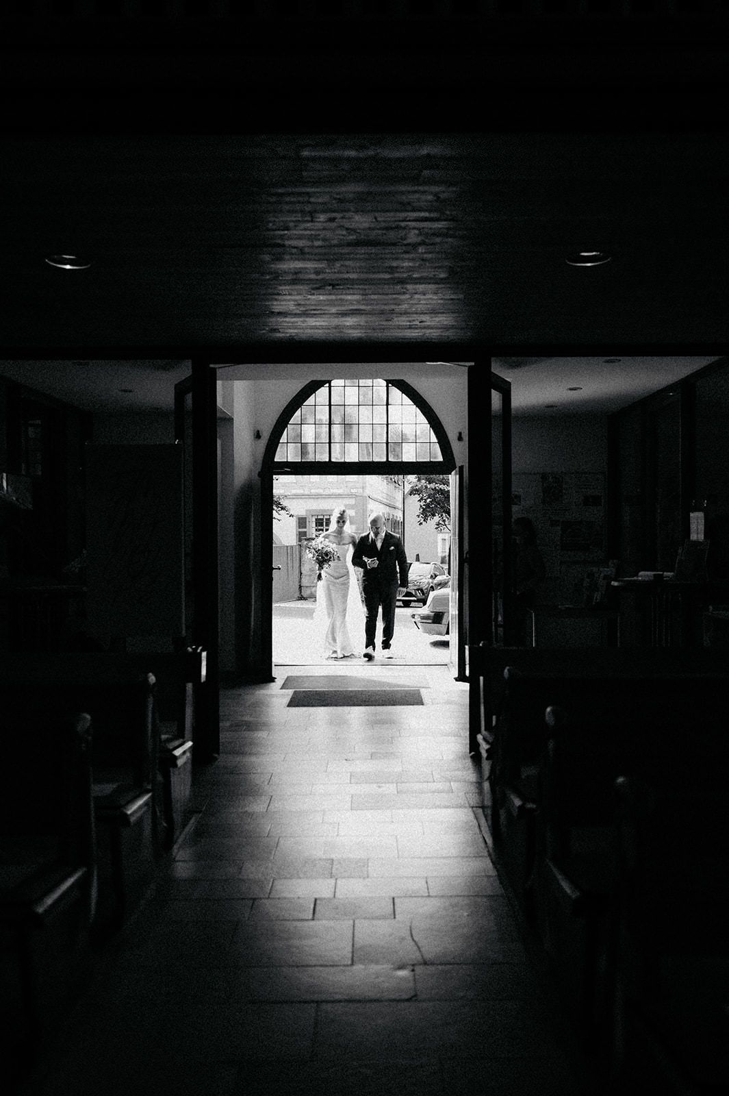 A silhouetted couple stands in an arched church doorway, framed by dark pews and dramatic lighting.