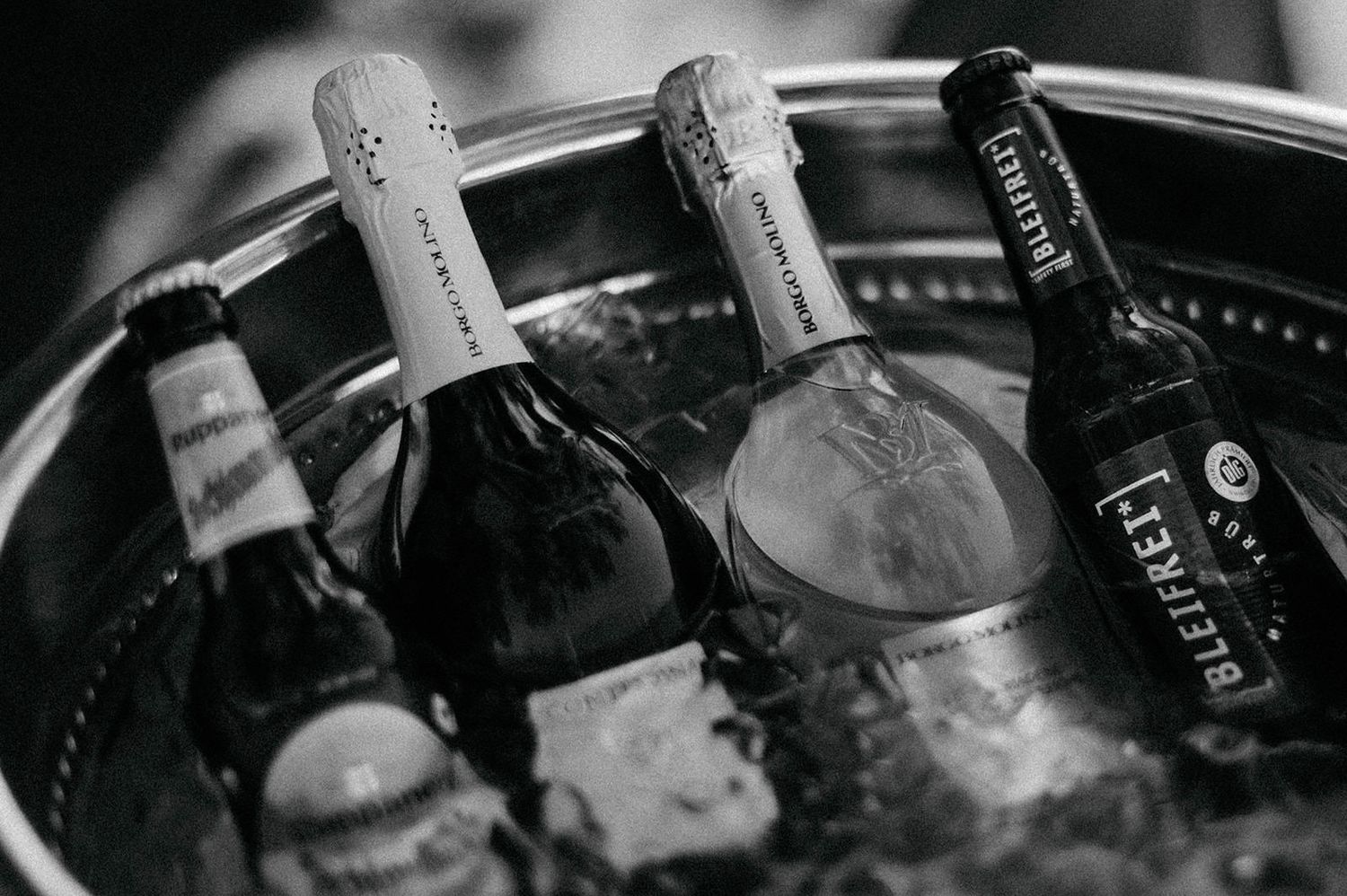 Black and white close-up of whiskey bottles on a bar shelf showing various spirits and glassware in low light.