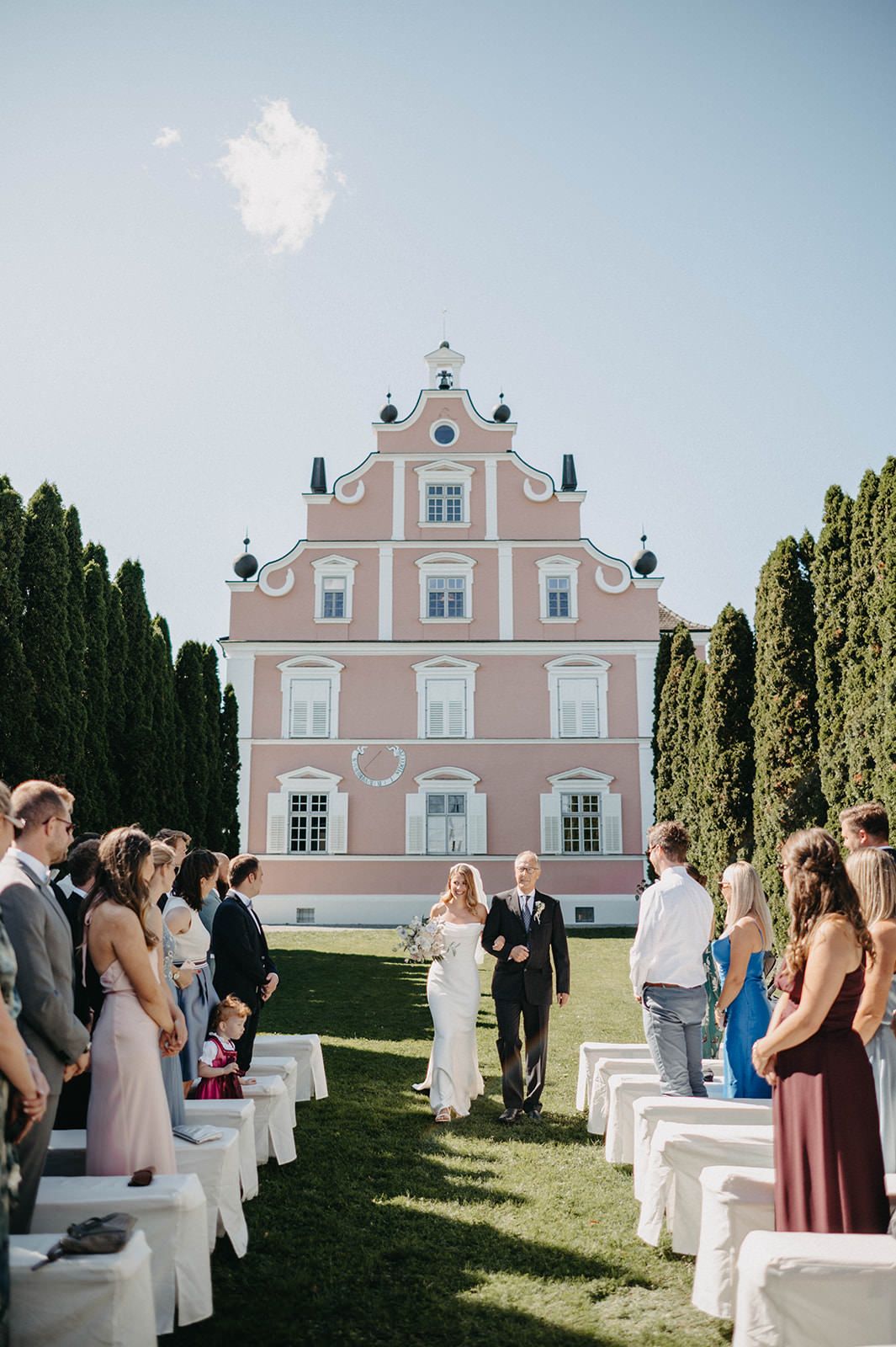 An outdoor wedding ceremony takes place in front of a pink European-style mansion with tall cypress trees on either side.