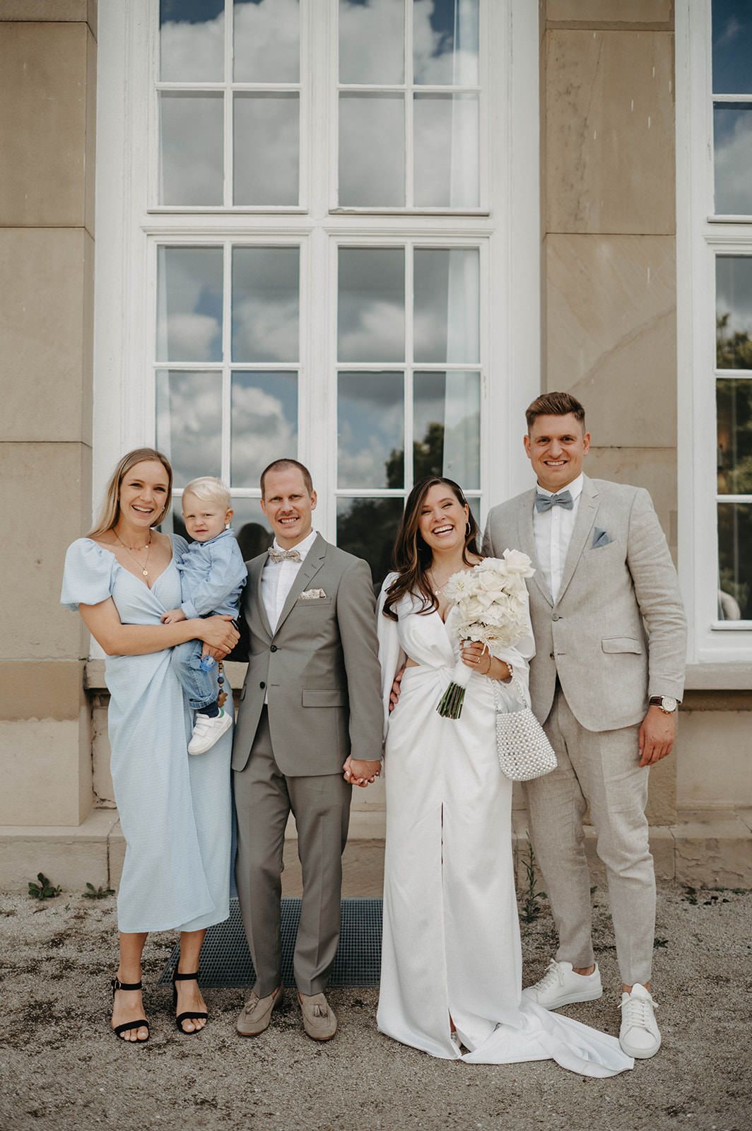 A wedding party poses outdoors in light blue and gray attire against a white building with large windows.