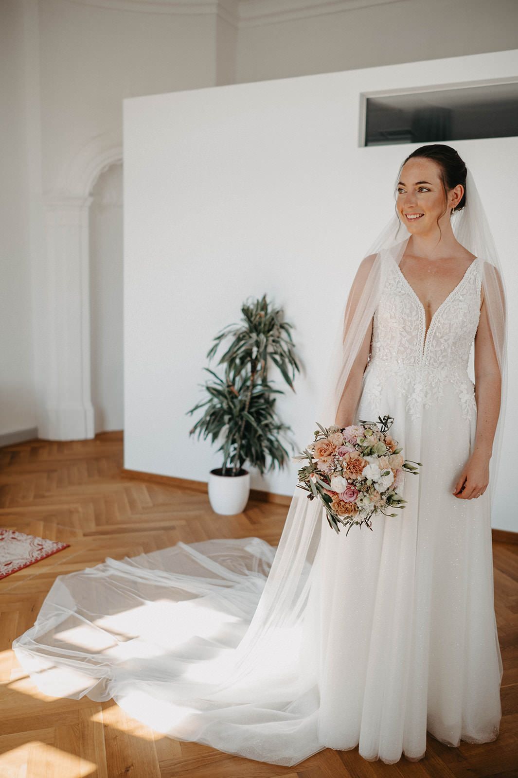 A bride in a flowing white wedding dress stands in a sunlit room with hardwood floors and a floral bouquet.