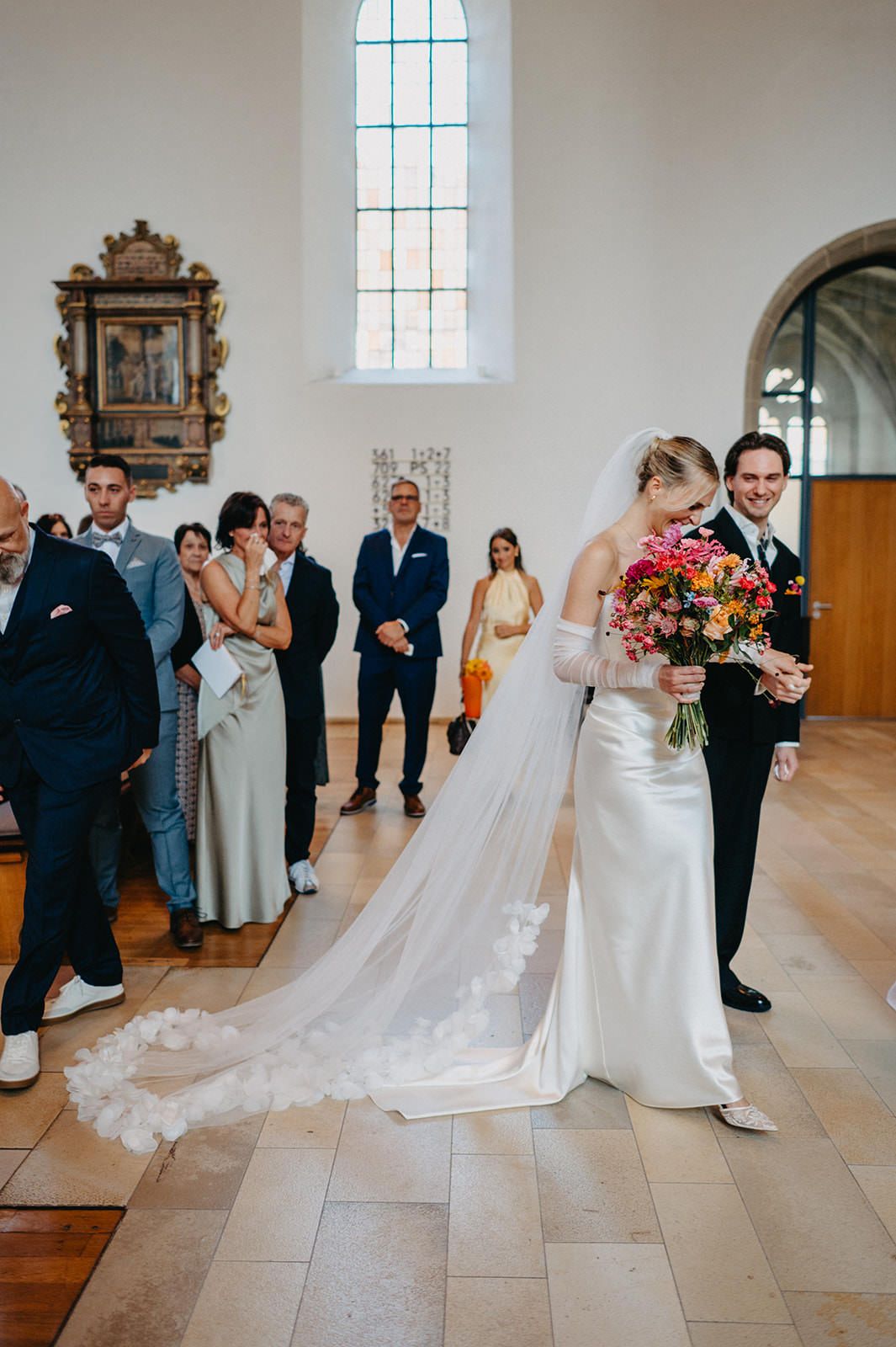 A wedding ceremony takes place in a bright church interior with stone floors and tall windows.