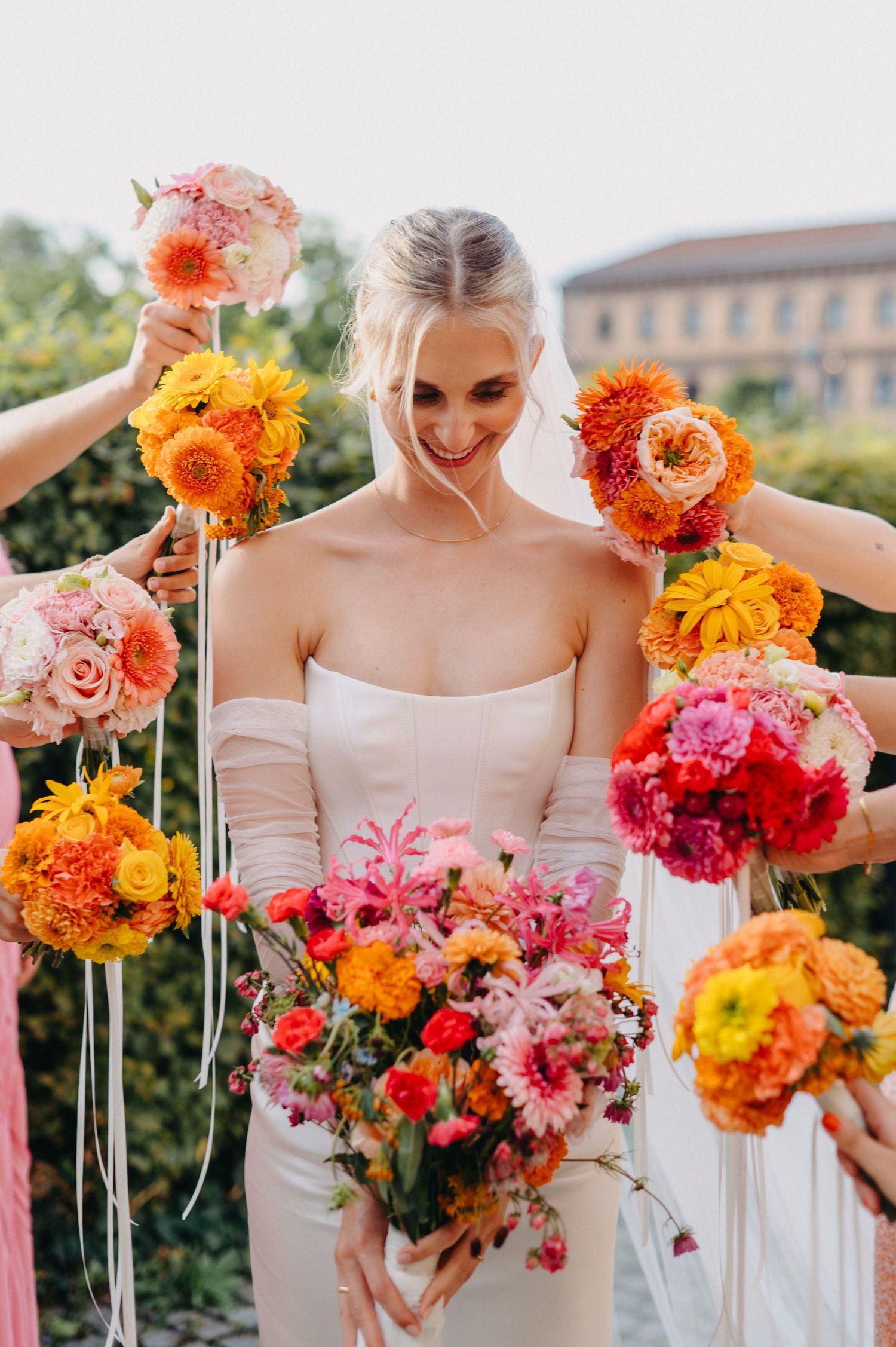 A bride in a white dress smiles while surrounded by vibrant orange, pink and yellow floral bouquets in a garden setting.