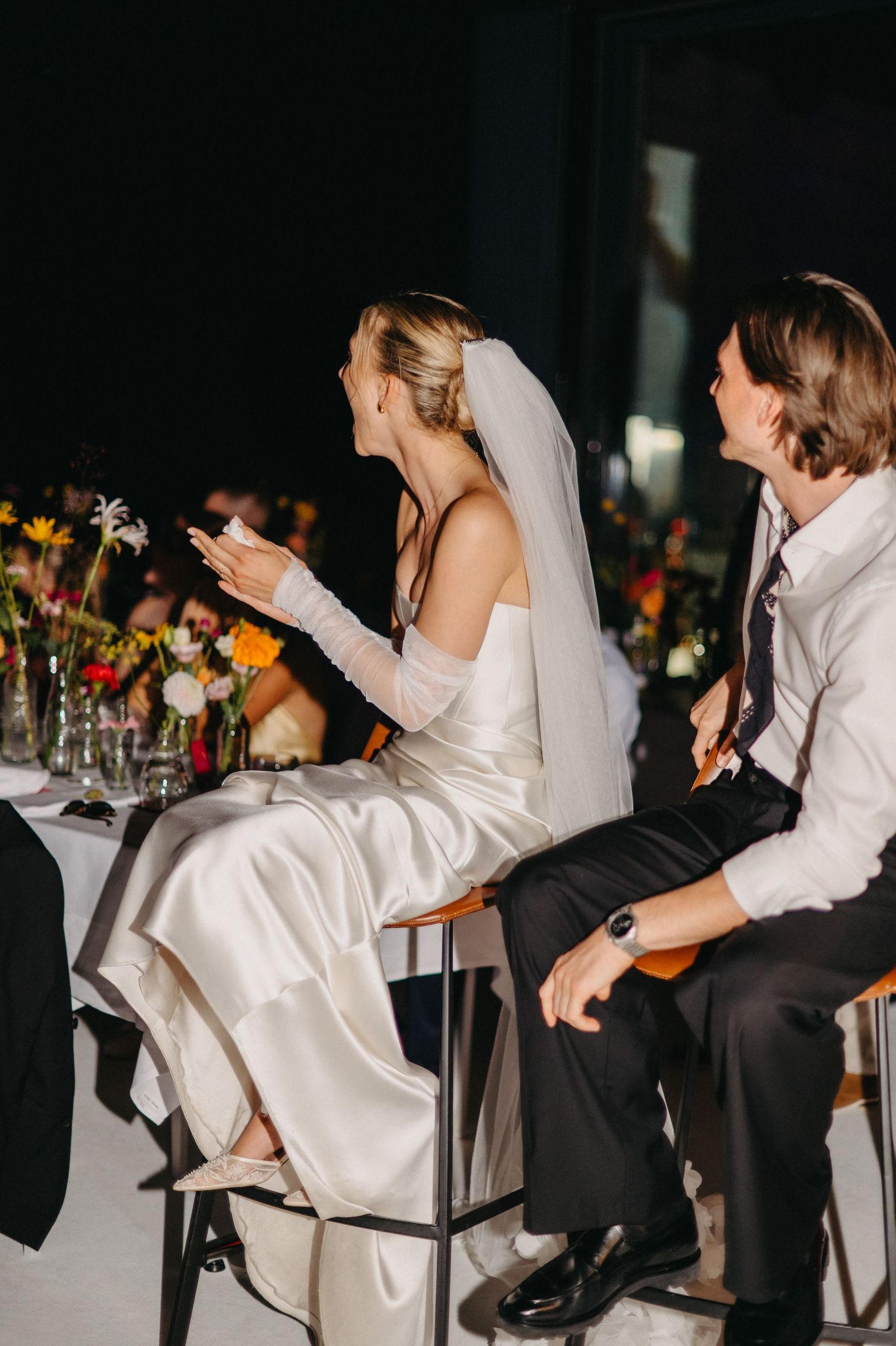 Wedding couple plays a fun game during reception while seated in chairs with floral arrangements in background.