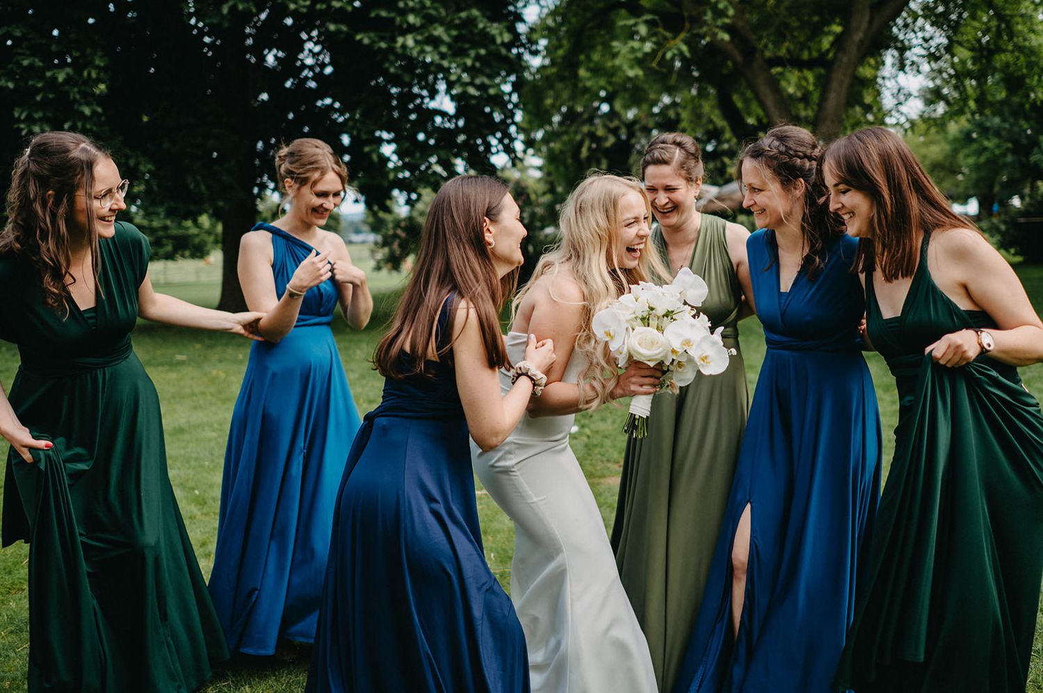 Bridal party in coordinating blue and green dresses share a candid laughing moment outdoors on a sunny day.